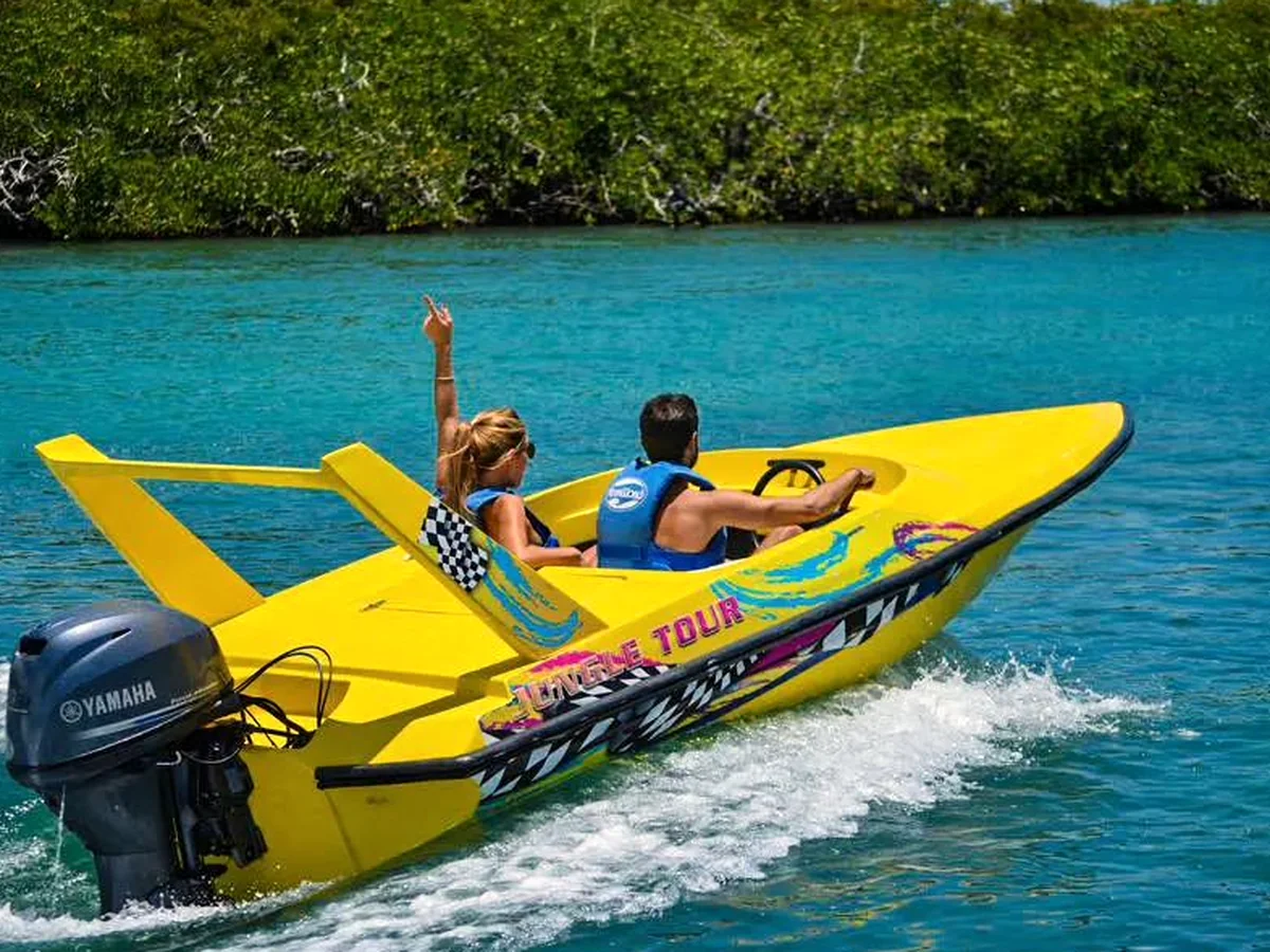A bright yellow two-seat speedboat cutting across the turquoise lagoon with a wall of mangrove behind it, the passenger's arm raised