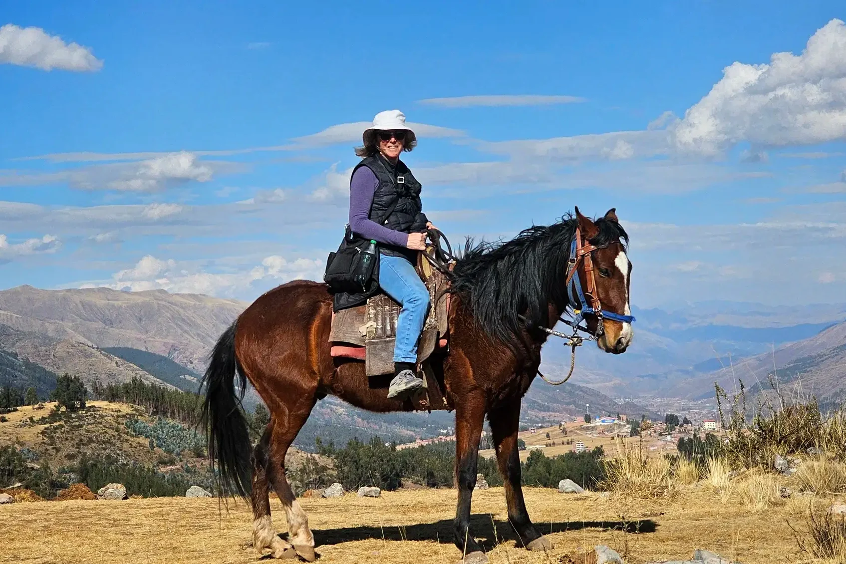 A rider in a white sun hat on a bay horse, standing on a dry ridge with the Cusco valley and the edge of the city far below