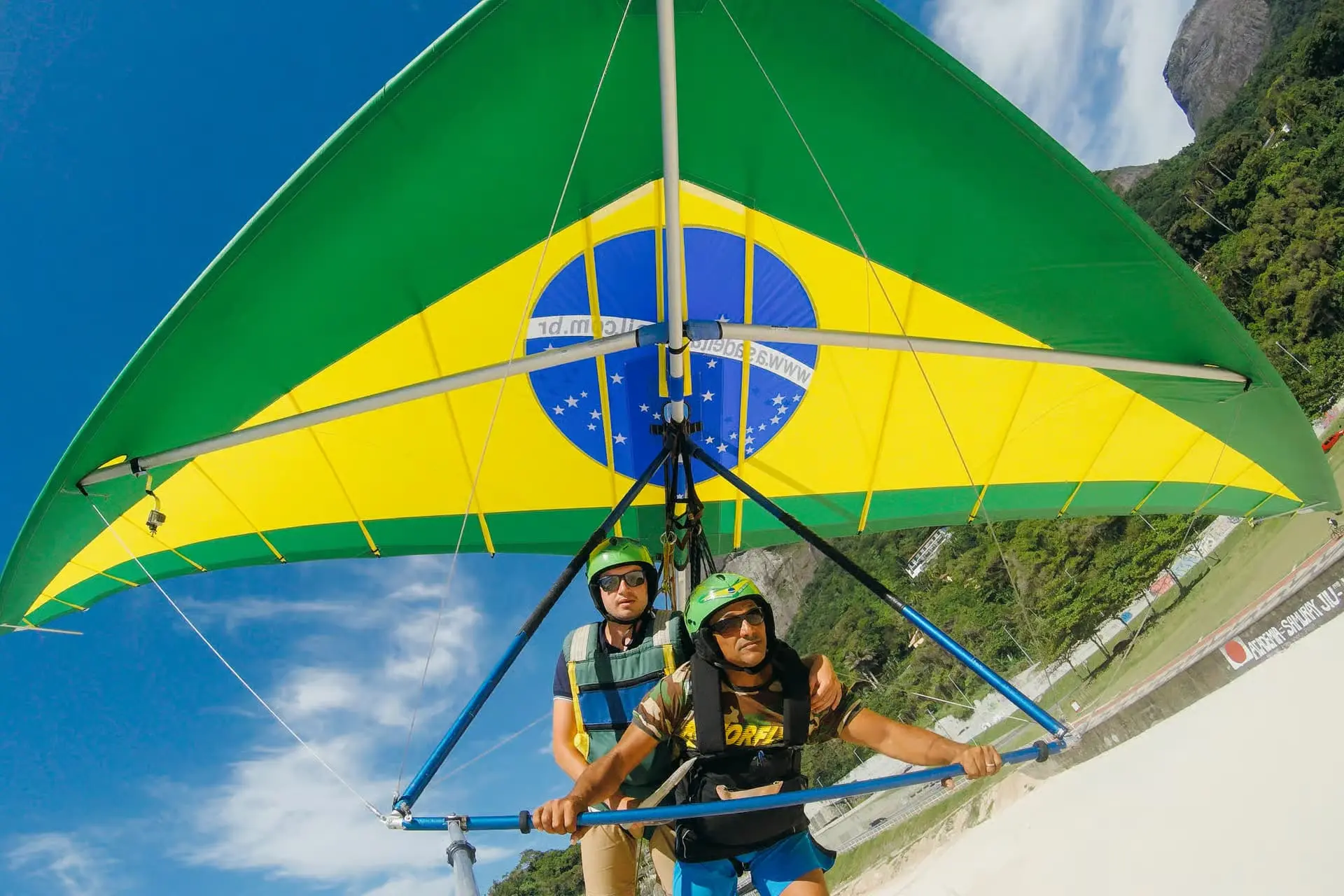 A tandem hang glider under a green and yellow wing painted as the Brazilian flag, the pilot and passenger holding the bar with forest and the beach road tilting away below