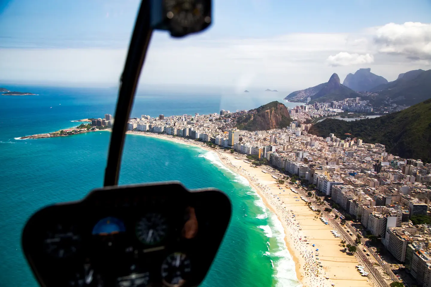 The curve of Copacabana seen past a helicopter's instrument panel, the beach and the city meeting the green water, with Dois Irmãos and Pedra da Gávea beyond