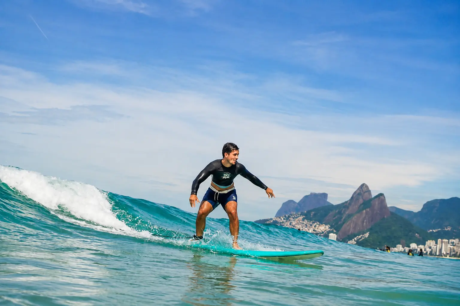 A beginner riding the white water on a soft surfboard at Arpoador, arms out and knees bent, the Two Brothers peaks and the far end of the beach behind