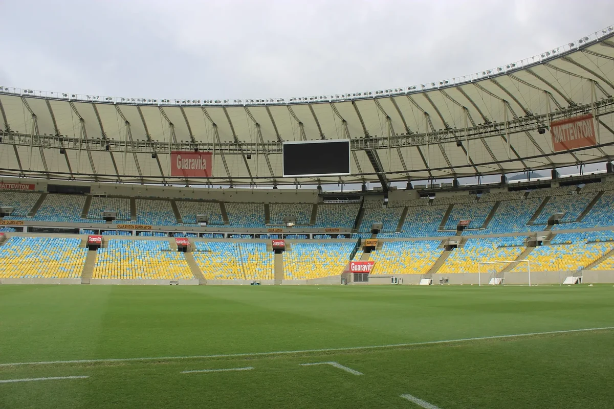 The empty bowl of the Maracanã from pitch level, blue and yellow seats rising in tiers under the ring of roof, the grass striped and unmarked