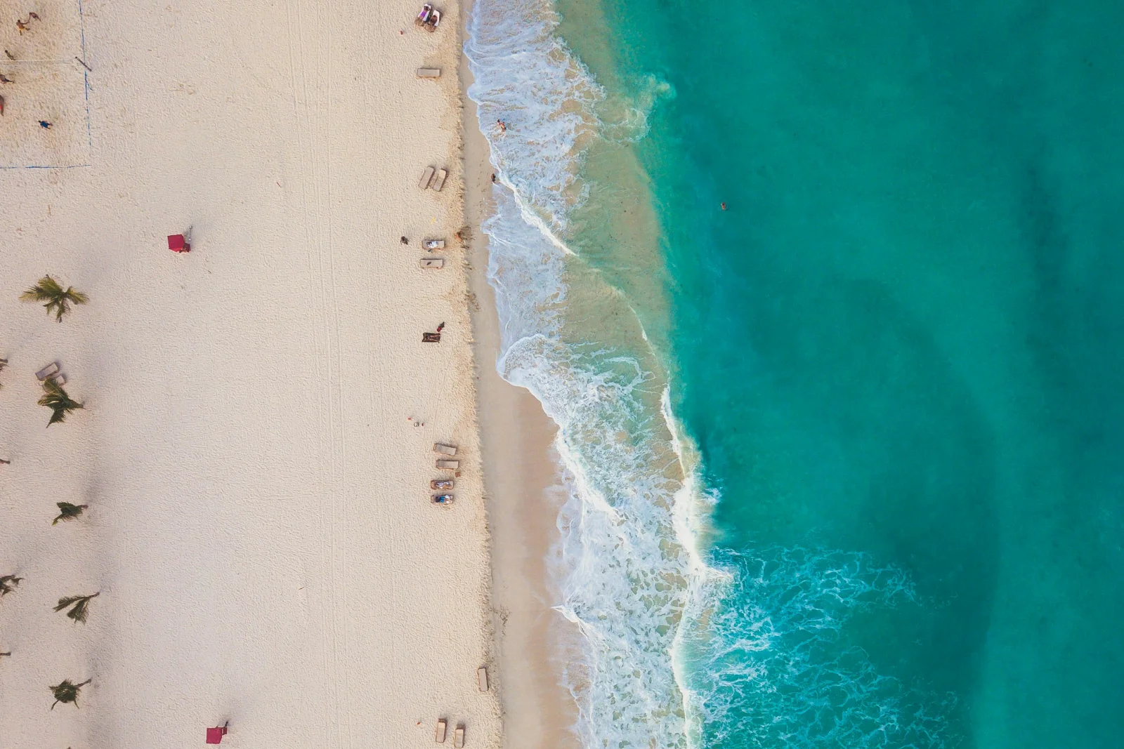 A Cancún shore from directly above, loungers and palm shade on the sand and surf running into turquoise water