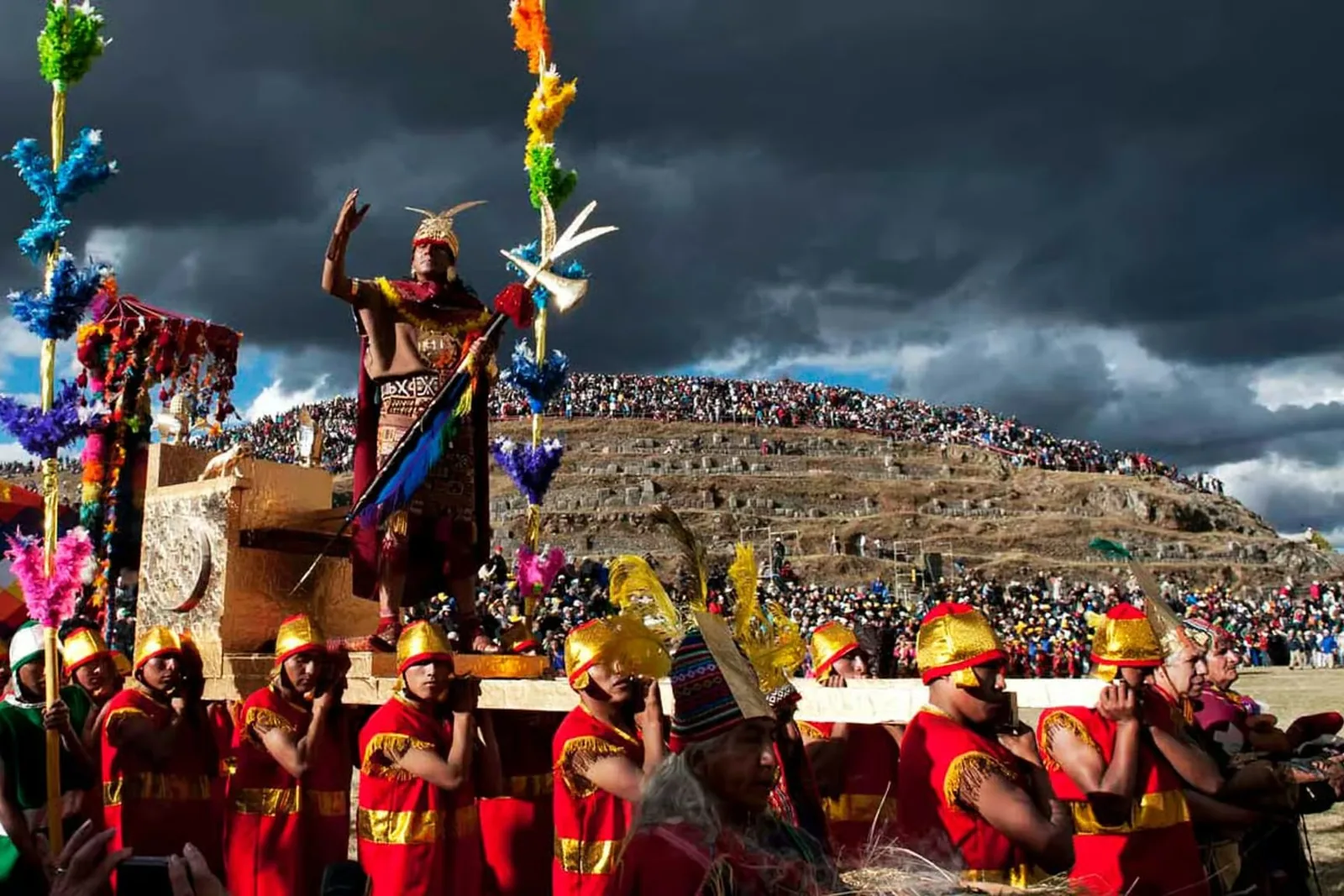 Ator como o Inca numa liteira dourada levada por homens de vermelho e ouro, com público no morro sob nuvens de tempestade