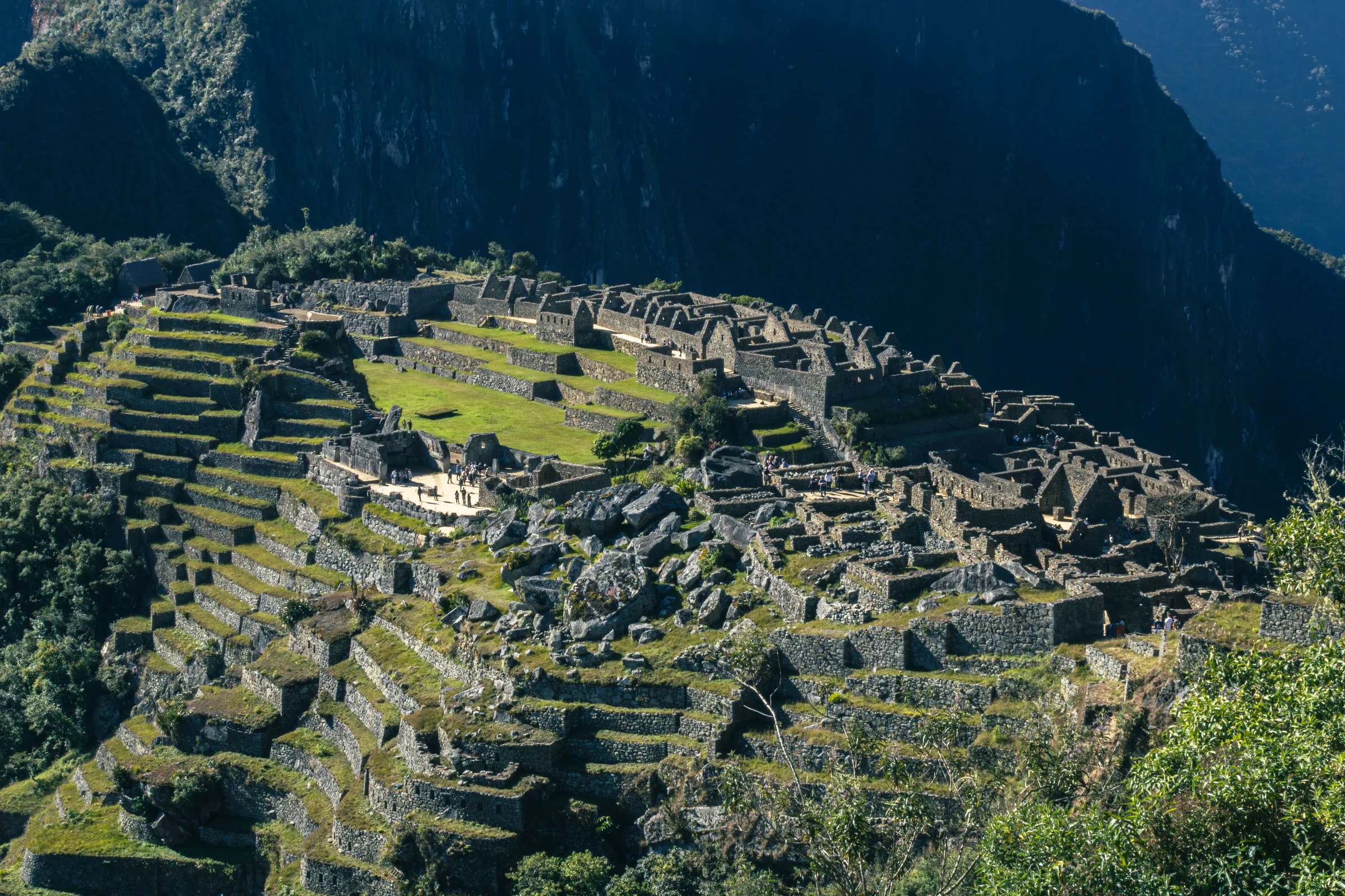 Os edifícios de pedra e os terraços verdes de Machu Picchu vistos do alto, com uma encosta escura de mata ao fundo