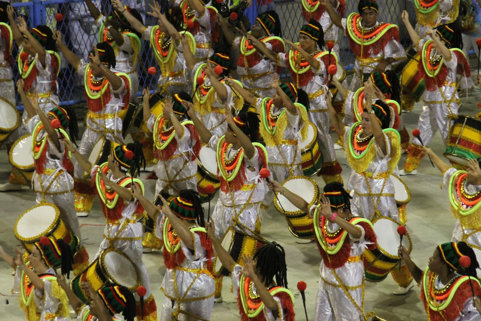Samba school drummers in white costumes with neon collars raise their arms among large drums, seen from above