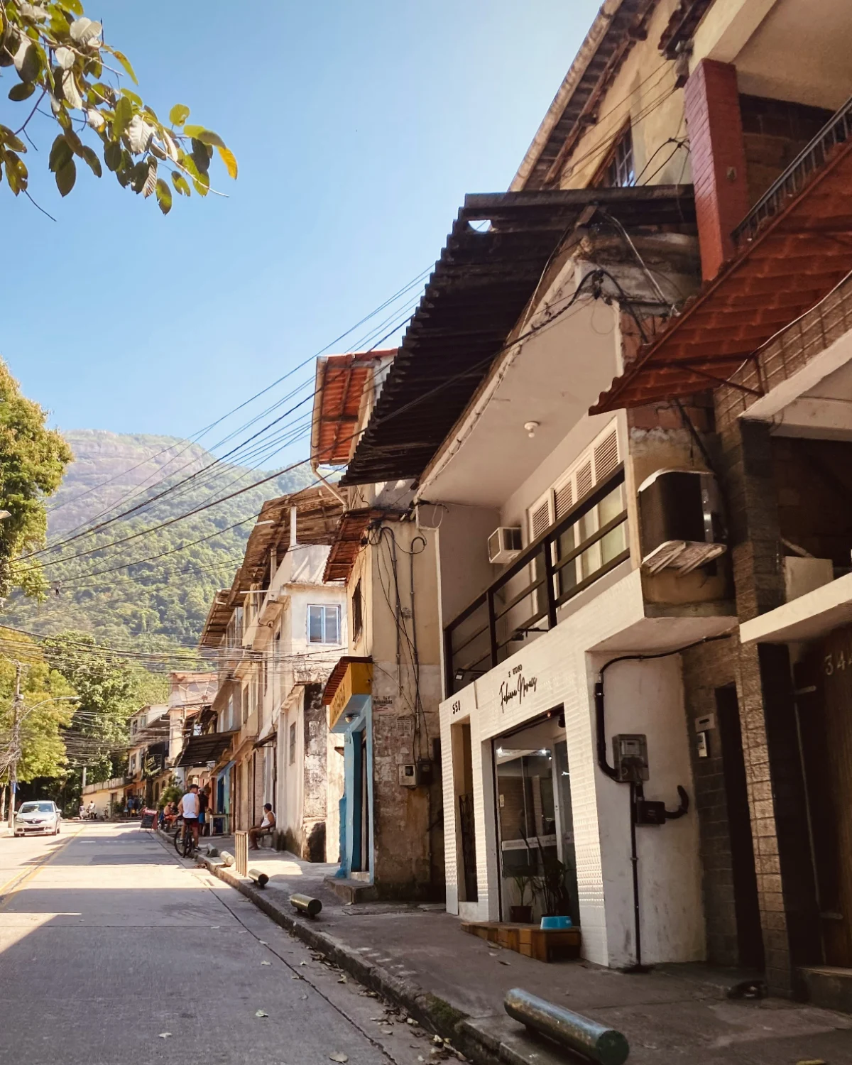 Calle residencial con tiendas y toldos, un ciclista y personas sentadas más adelante, y una colina boscosa al fondo