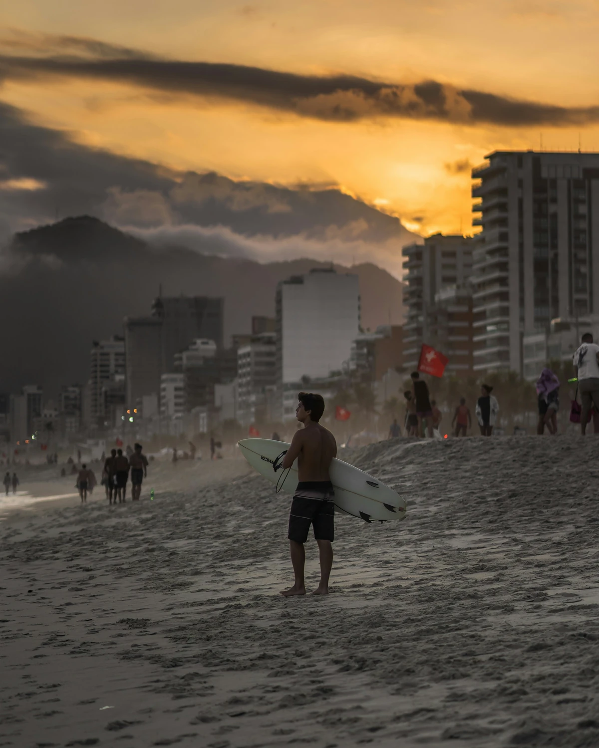 A surfer stands on the sand at dusk with a board under one arm, apartment blocks and a cloud-covered peak behind him