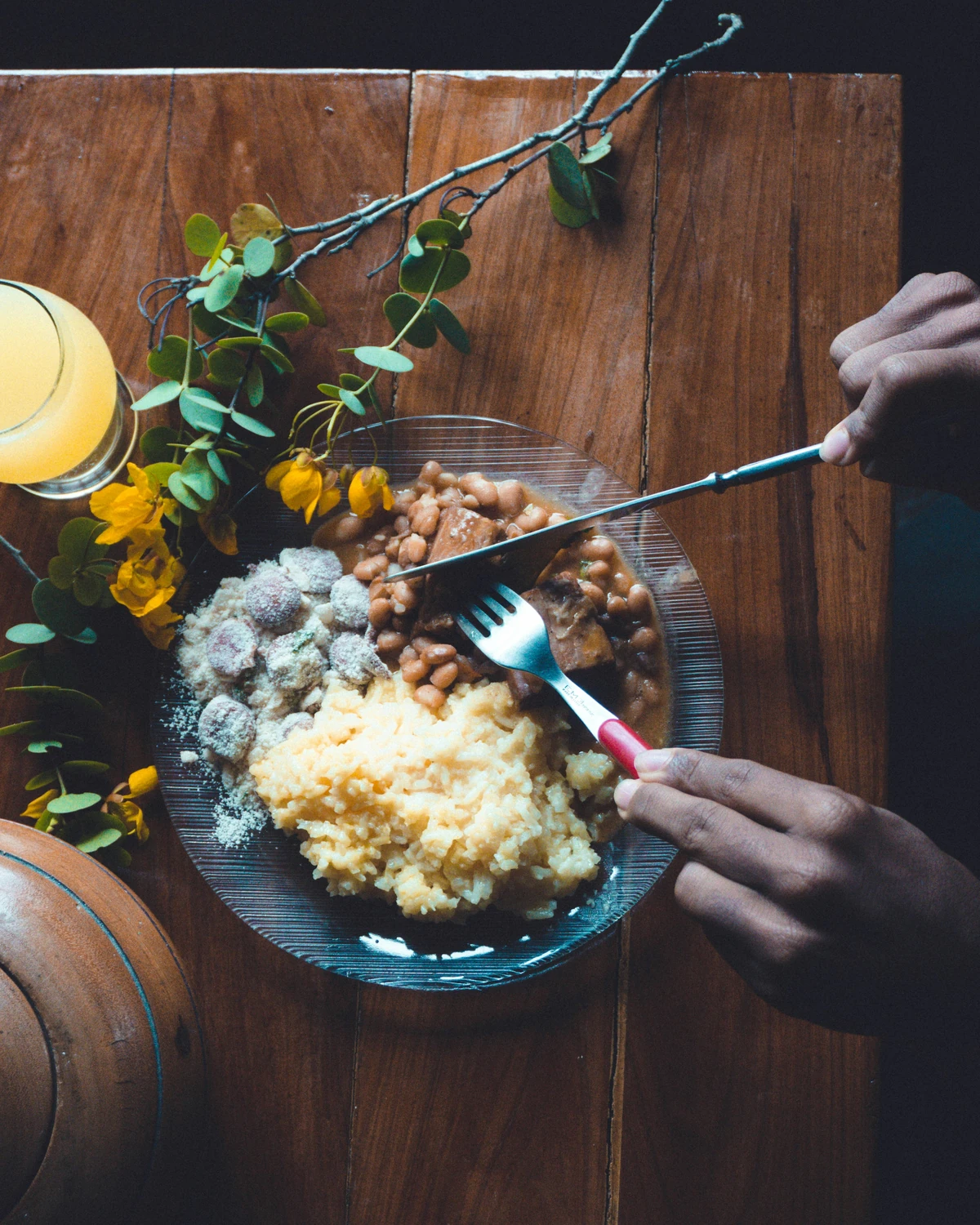 Two hands with a knife and fork over a plate of rice, beans and farofa on a wooden table, a glass of juice beside it