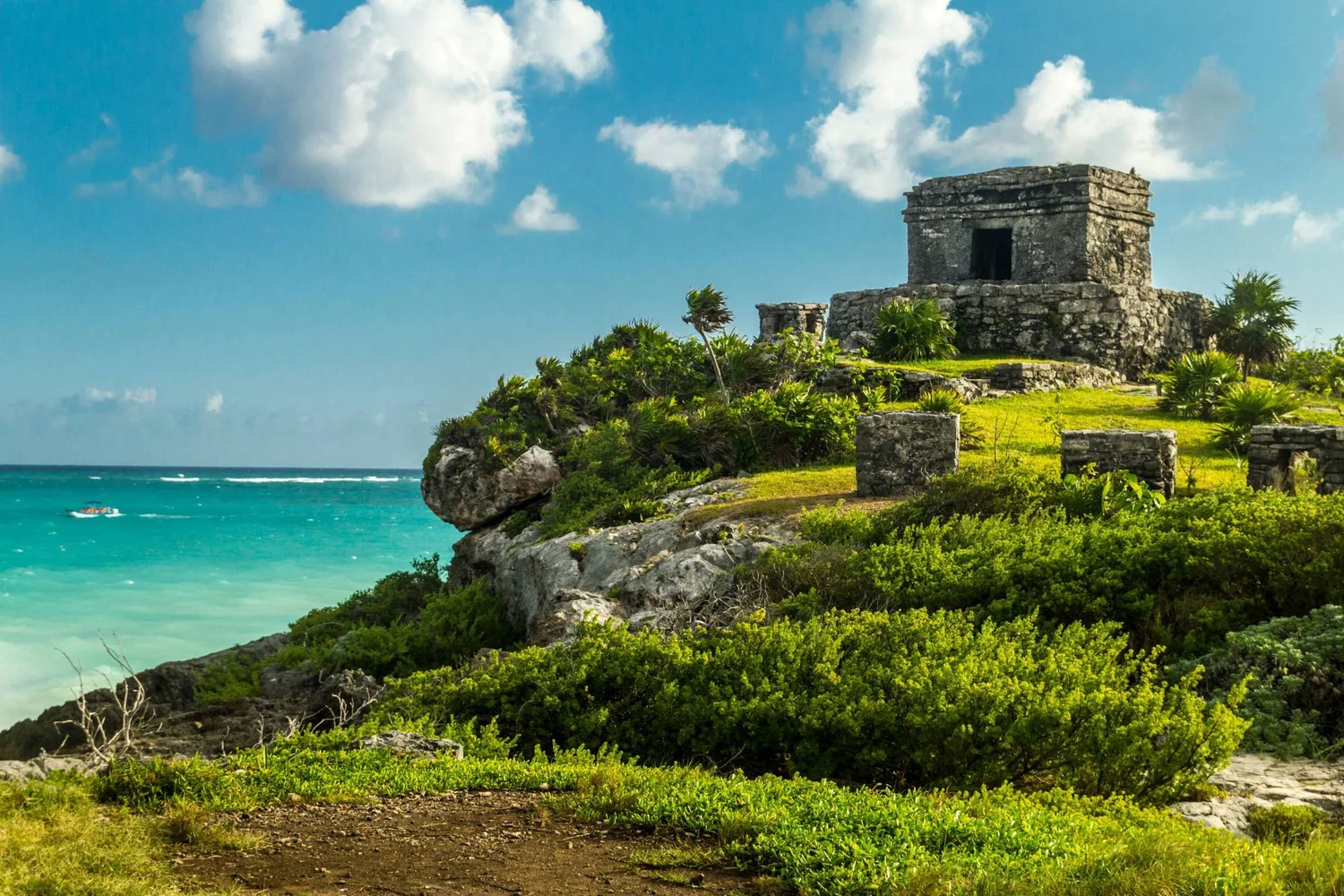 A small stone temple of Tulum on a green rise above the turquoise Caribbean, a boat on the water below