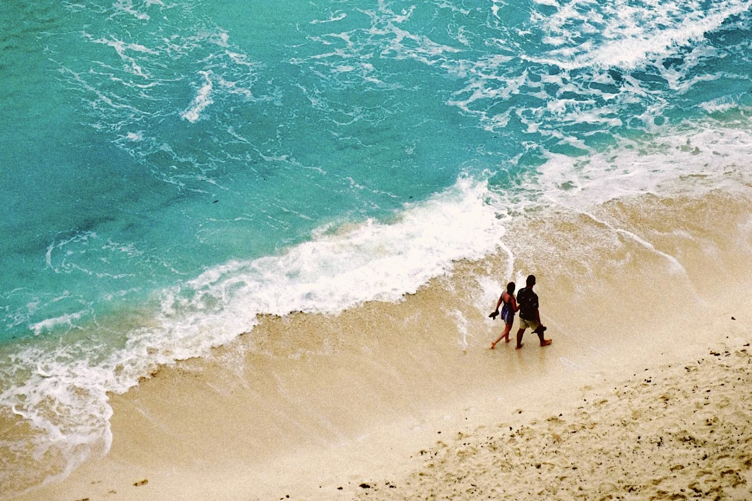 Two people walking along the edge of the surf on a pale sand beach, seen from above, turquoise water breaking beside them