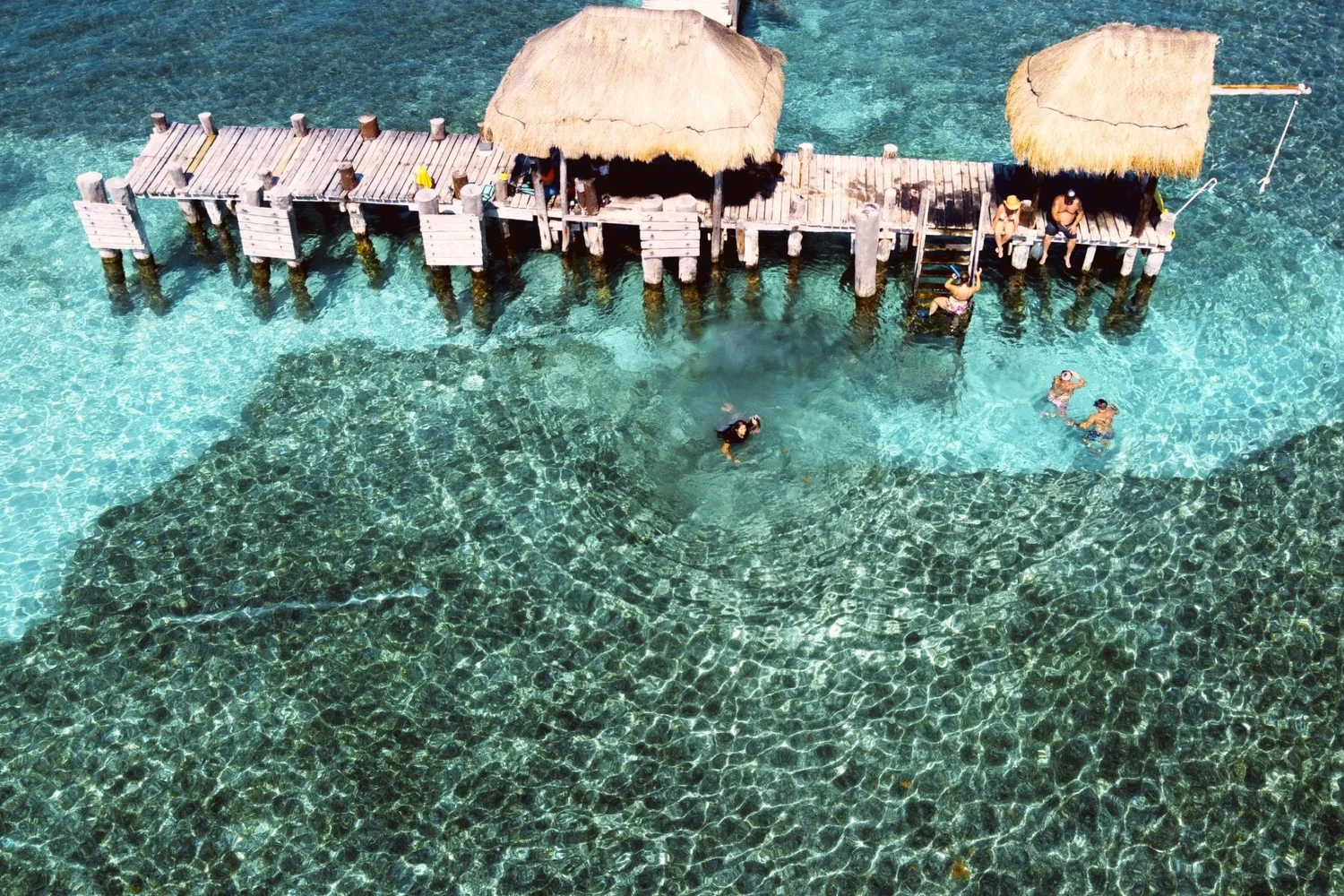 A wooden pier with two thatched palapas over clear turquoise water at Isla Mujeres, seen from above, swimmers below