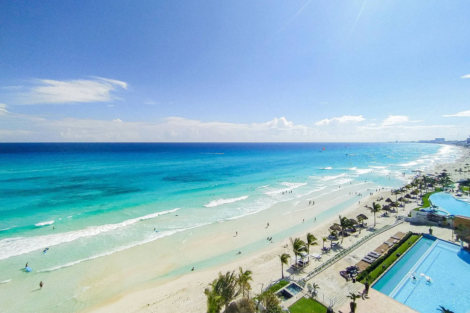 The Hotel Zone beach from a high floor, turquoise water and white surf along pale sand, palapas and palms behind it, hotel pools in the foreground and the strip curving away to the horizon