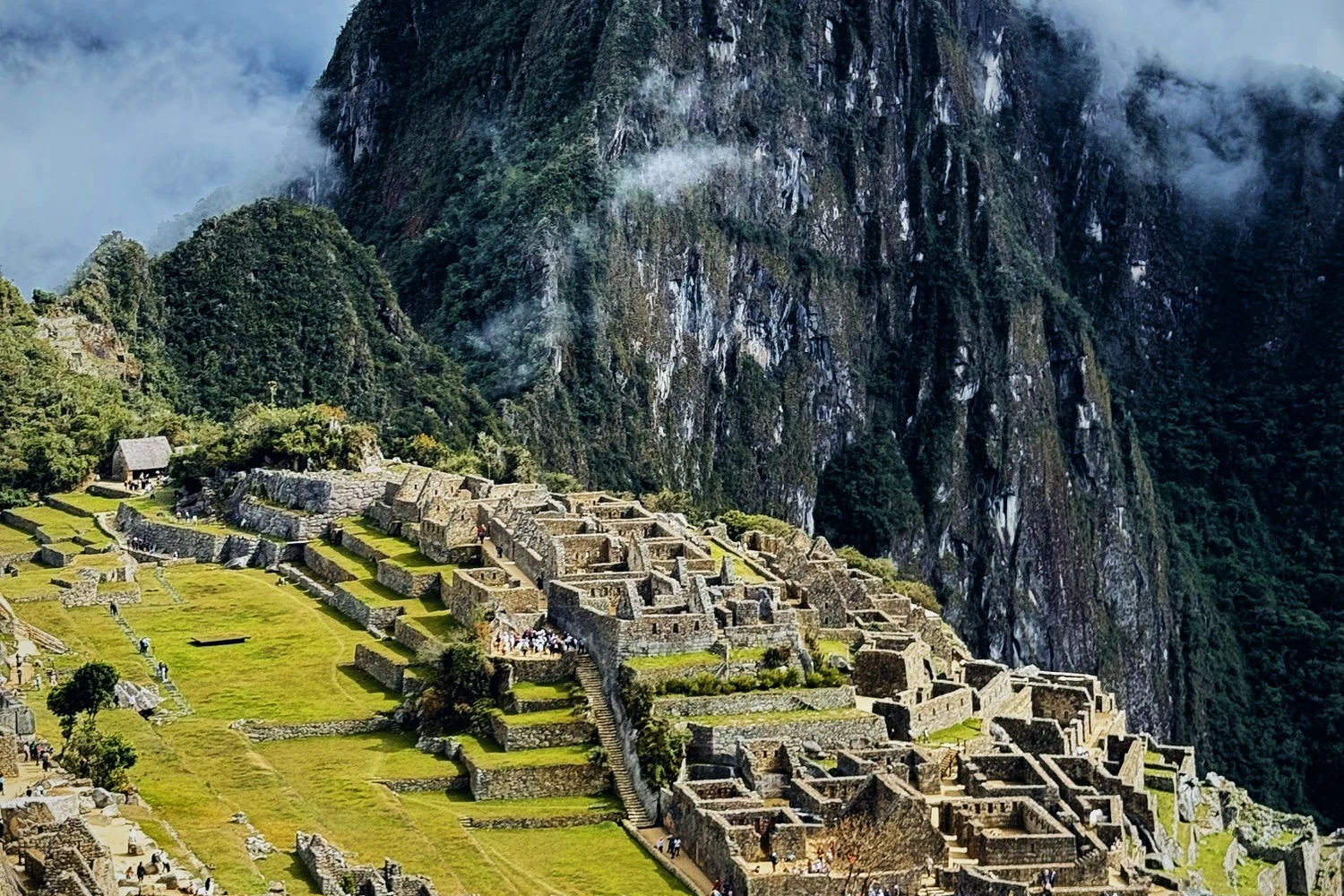 The stone terraces and houses of Machu Picchu on a green ridge below the steep, cloud-touched face of Huayna Picchu