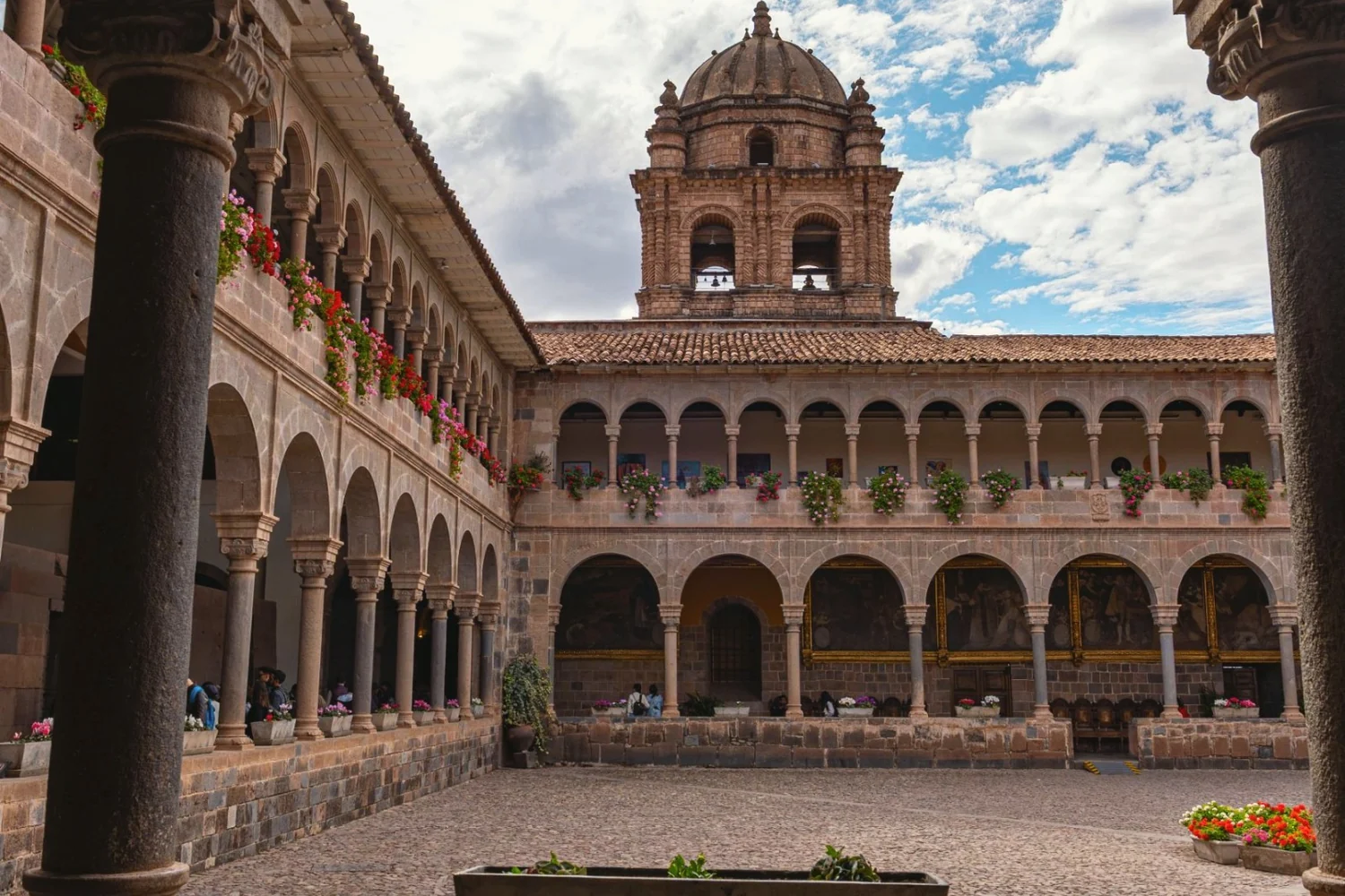 Stone arches of the Qorikancha cloister framing a courtyard, the bell tower of Santo Domingo above under a blue sky with clouds