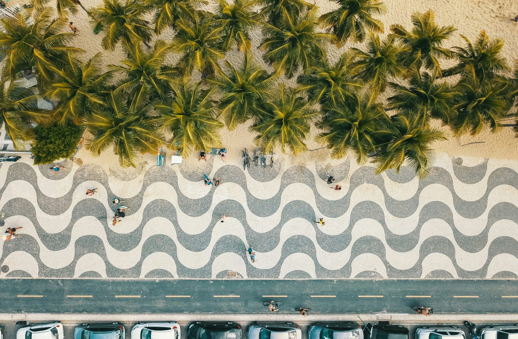 The black and white wave mosaic of the Copacabana promenade seen from directly above, with palms and a few walkers between the sand and the road