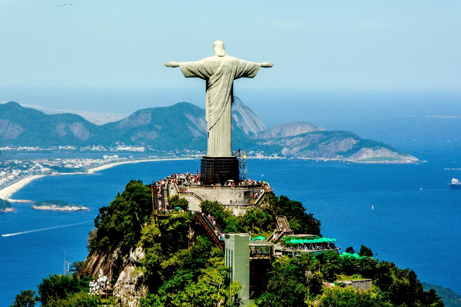 El Cristo Redentor en la cima del Corcovado, bajo una luz clara, con los brazos abiertos sobre el bosque y las playas y el mar abierto muy abajo