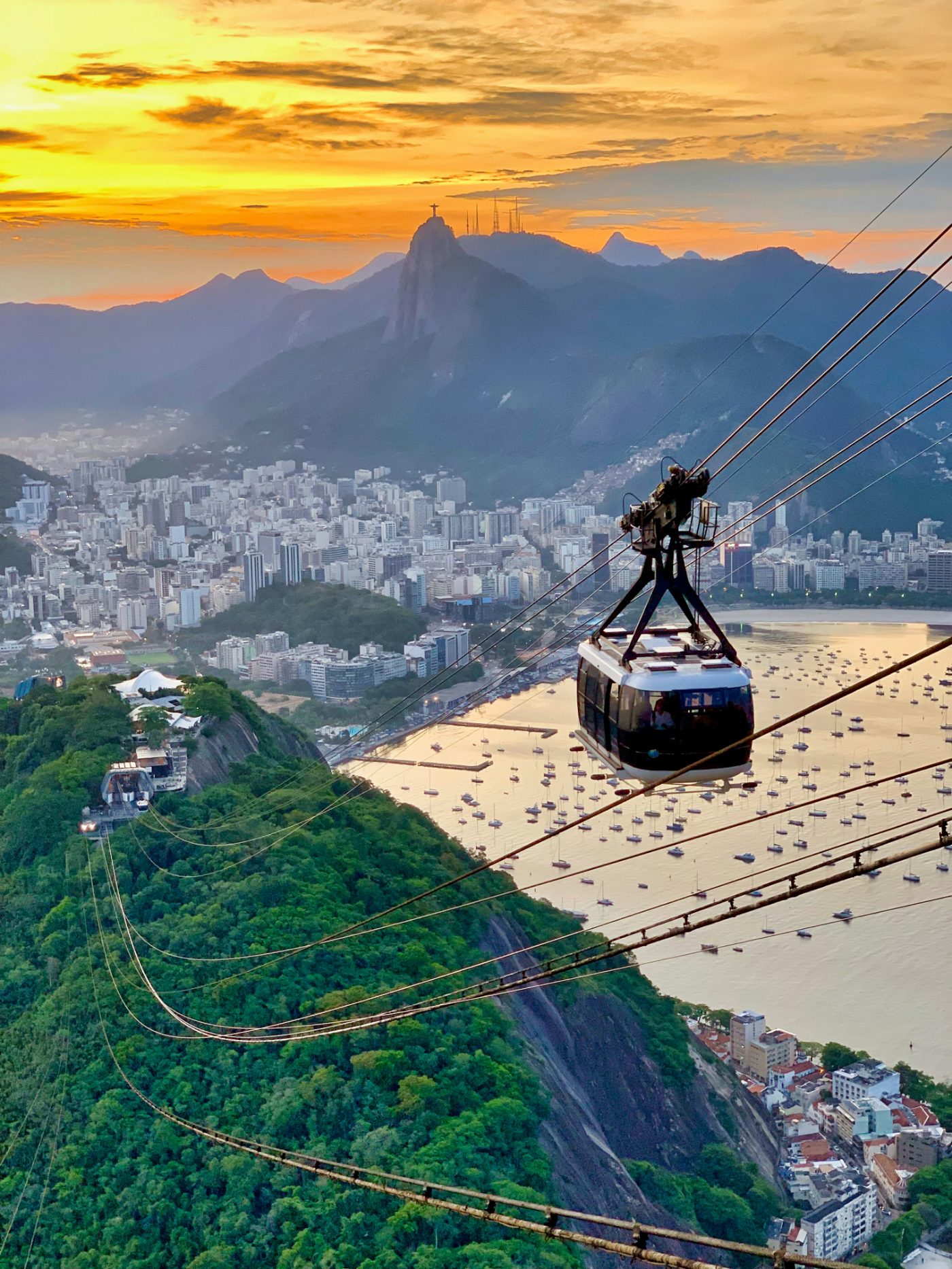 A cable car riding its lines toward Sugarloaf at sunset, with Christ the Redeemer on the far ridge and the moored boats of Botafogo Bay far below