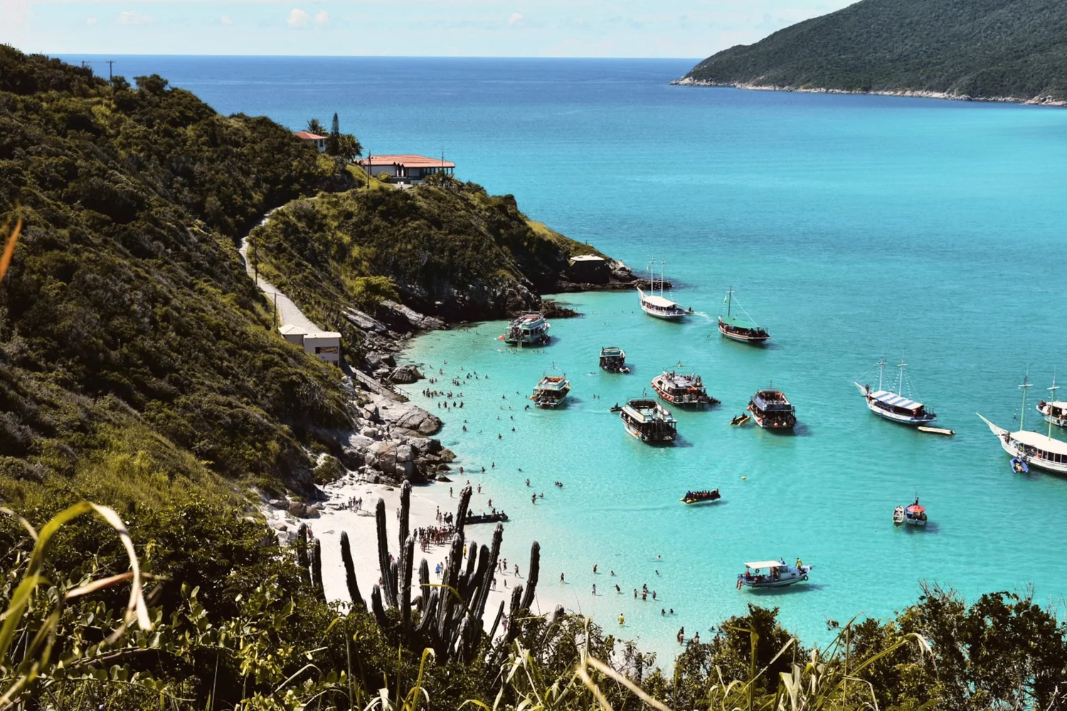 A turquoise cove at Arraial do Cabo below a green hillside with cactus, boats anchored in the clear water and a path down to the sand