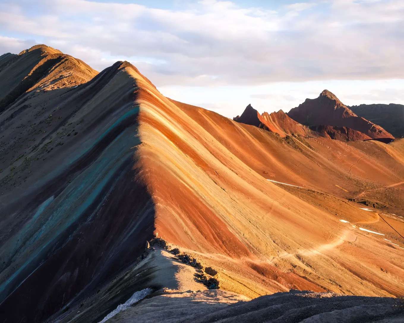 The striped ridge of Rainbow Mountain in low sun, with red peaks behind it