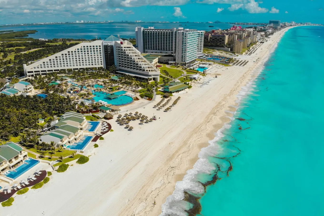 Resort hotels along the white sand of the Hotel Zone from above, with the turquoise sea on one side and Nichupté Lagoon behind