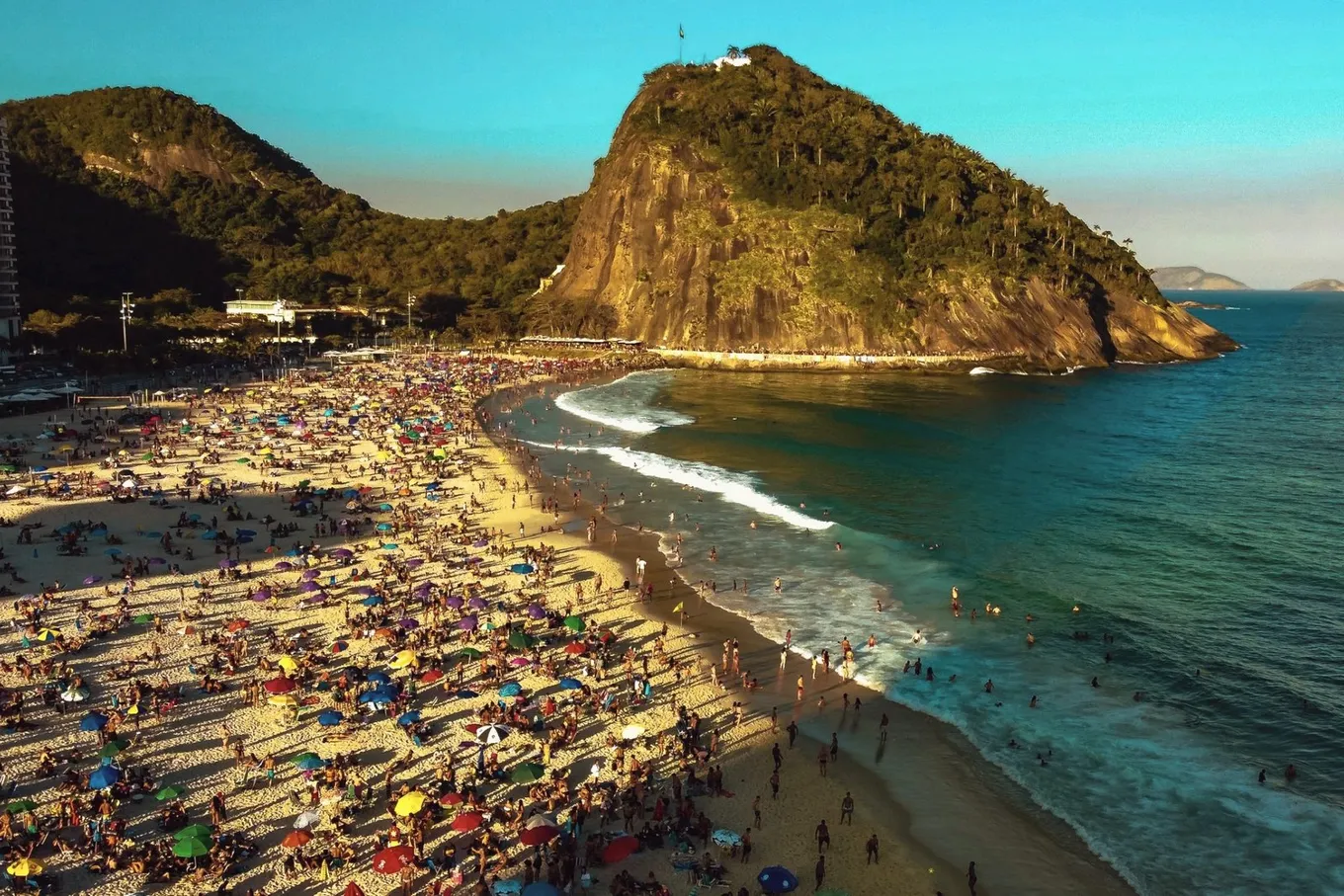Leme beach from above in late afternoon light, crowded with umbrellas below a forested headland