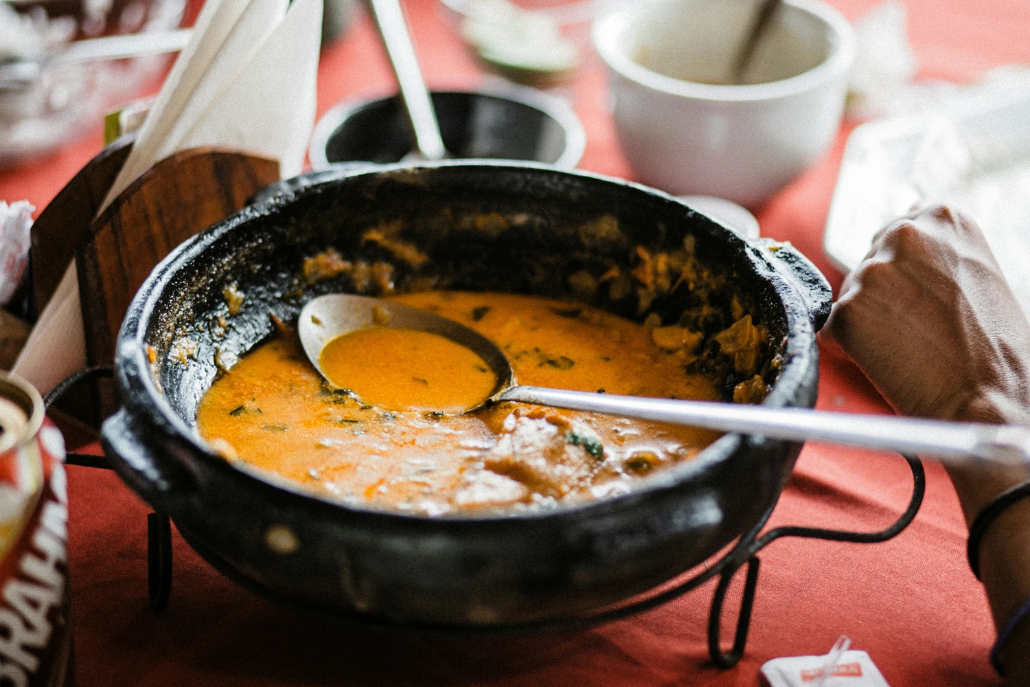 A black clay pot of moqueca on an iron stand over a red tablecloth, a serving spoon resting in the stew and a hand at the edge of the frame