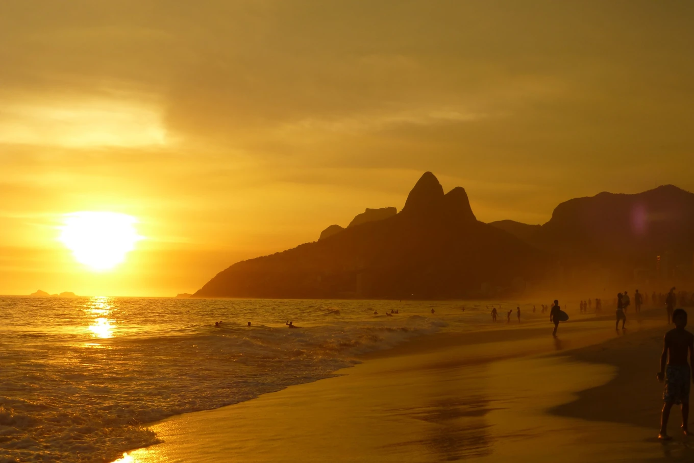 A beach at sunset, the low sun laying a path of light across the water, small figures on the wet sand, and a twin peak in silhouette down the shore