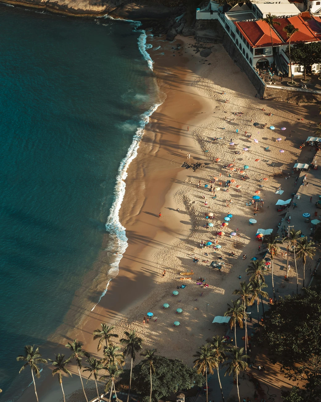 A curved beach seen from high above, colored umbrellas and long shadows across the sand, palms along its edge and rock at the far end