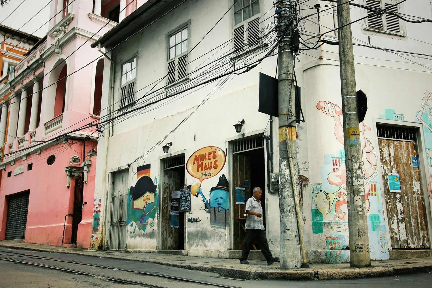 A street corner with tram rails in the road, painted murals on peeling plaster and one man walking past an open doorway